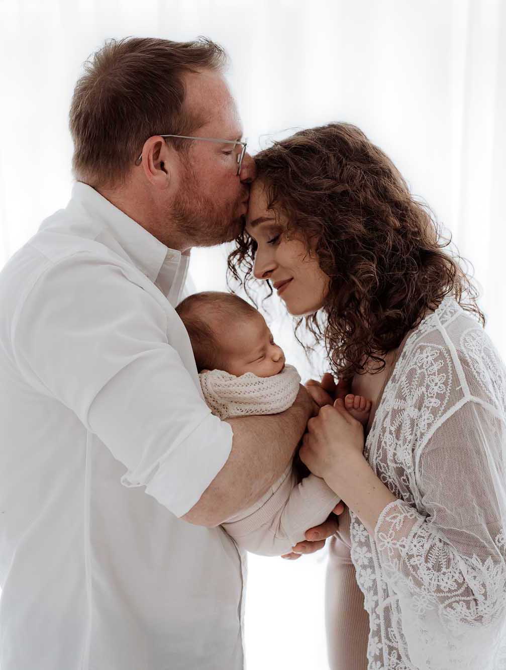 Mama und Papa stehen, er küsst sie auf die Stirn und sie halten ihr Baby zwischen sich auf dem Arm
