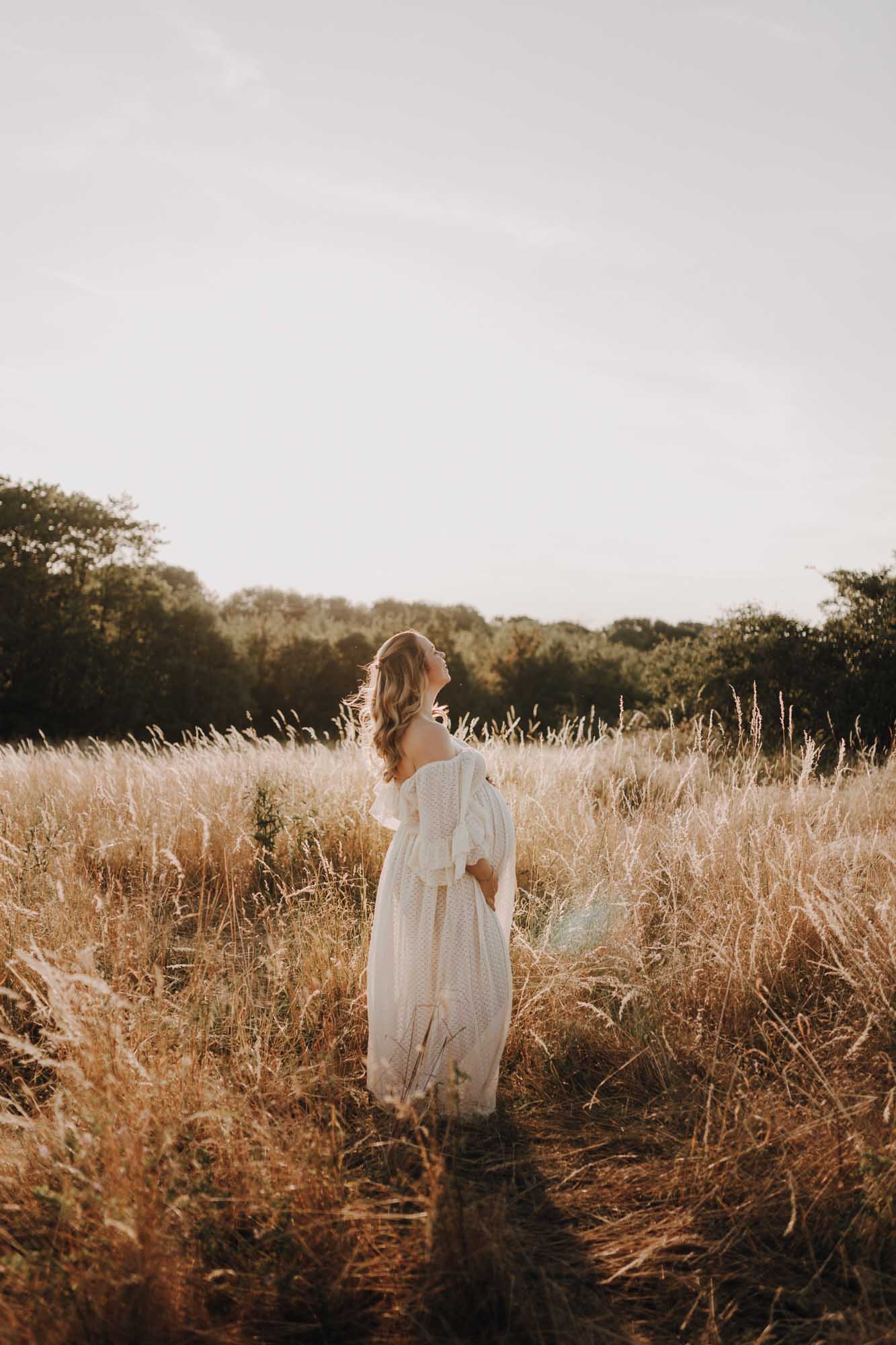 eine schwangere steht zwischen hohen Gräsern in der Abendsonne