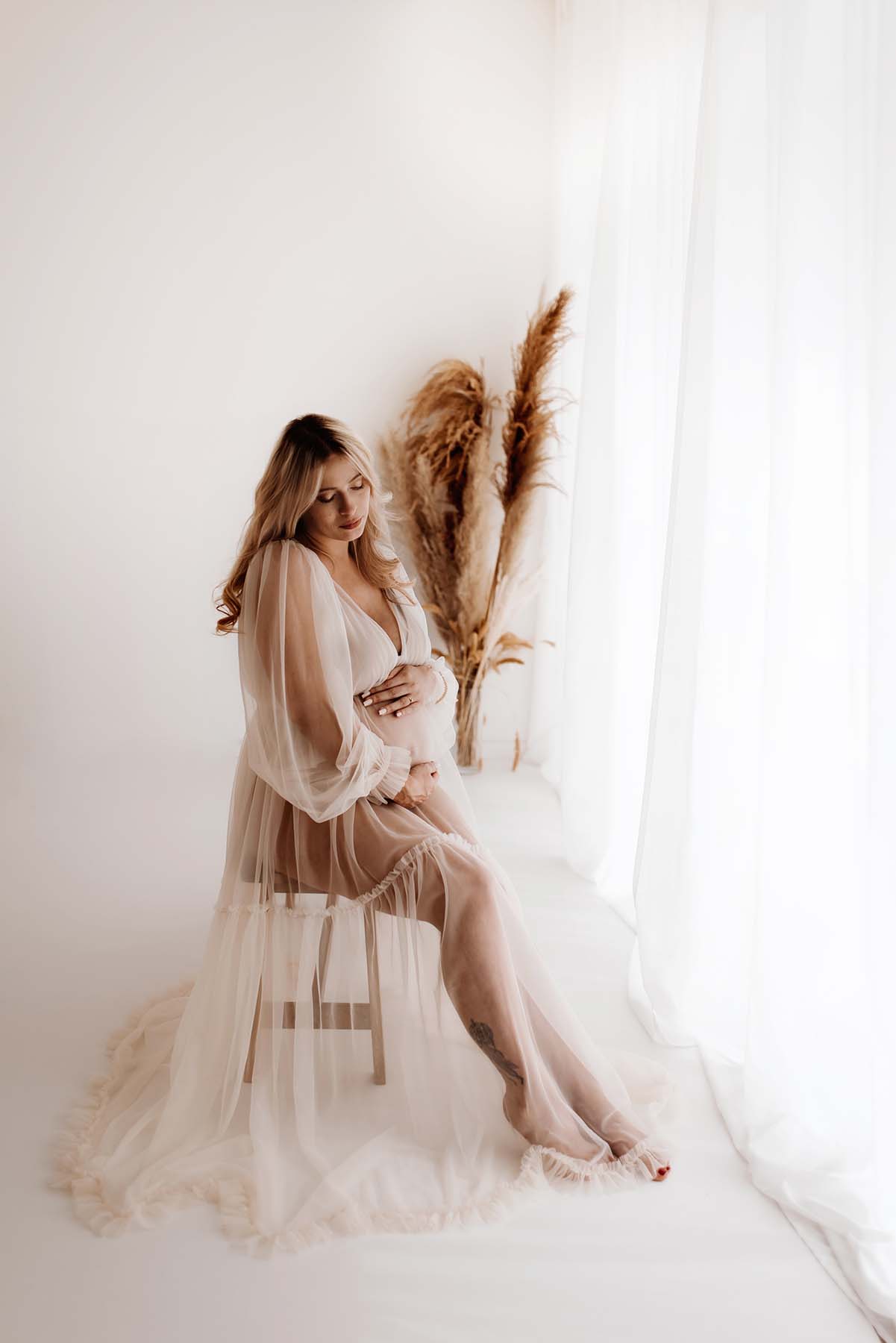 eine schwangere sitzt im Fotostudio auf einem weißen Stuhl mit pampasgras im Hintergrund